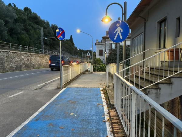 Fine pista ciclabile blu vicino strada principale. Un segnale indica la fine del percorso ciclo-pedonale, l’altro l’inizio del tratto pedonale.